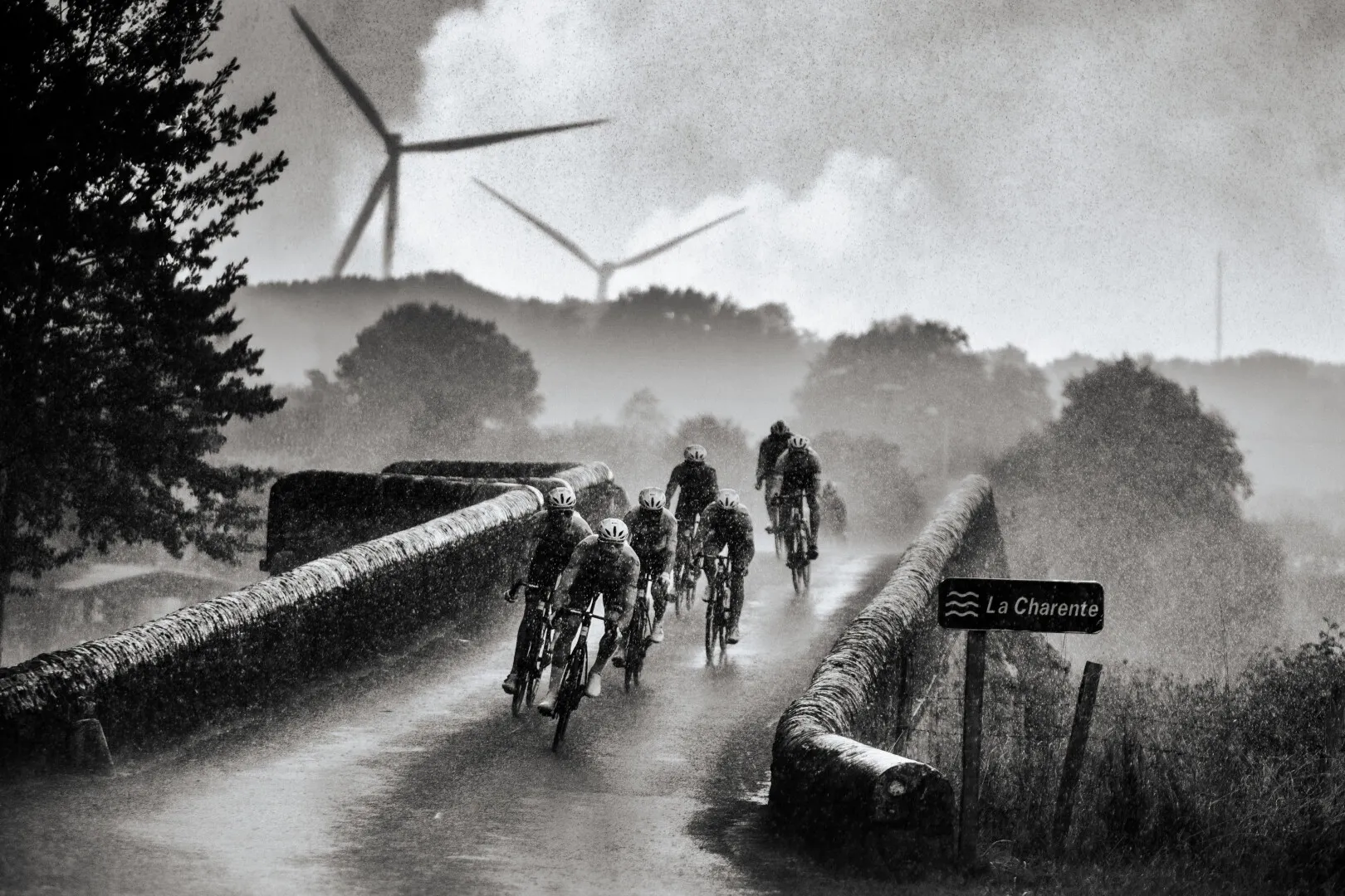 Cyclists riding in rainy weather on a narrow stone bridge over La Charente river with wind turbines and trees in the misty background.