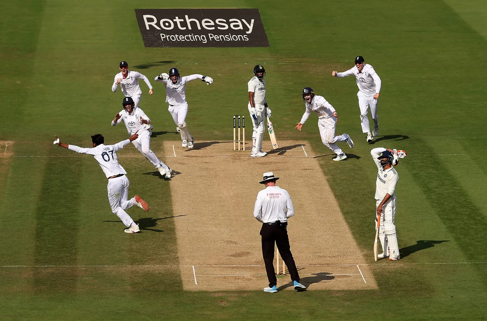 Cricket players in white uniforms celebrating a wicket on the pitch while the batsman looks on and the umpire watches.