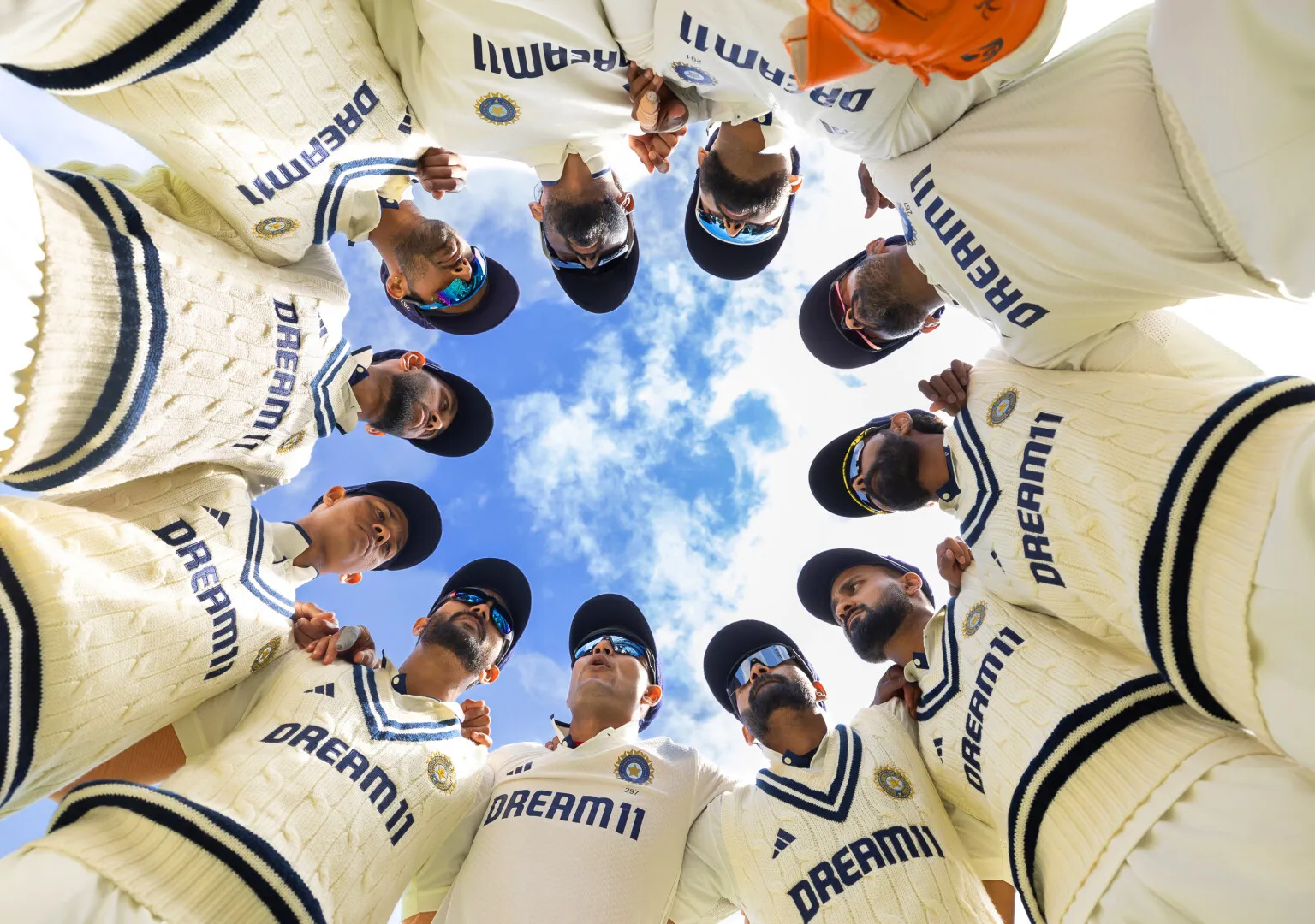 Indian cricket team players in white jerseys with Dream11 logos standing in a huddle under a blue sky.