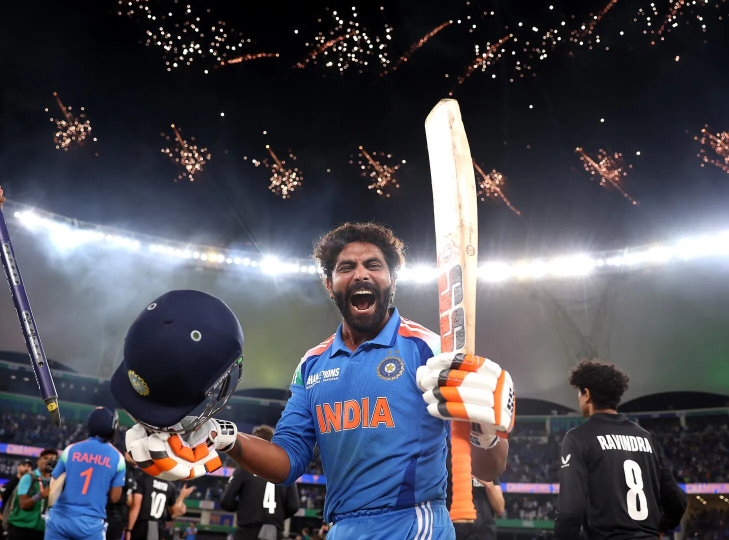 Indian cricket player celebrating with bat and helmet raised under stadium lights and fireworks.