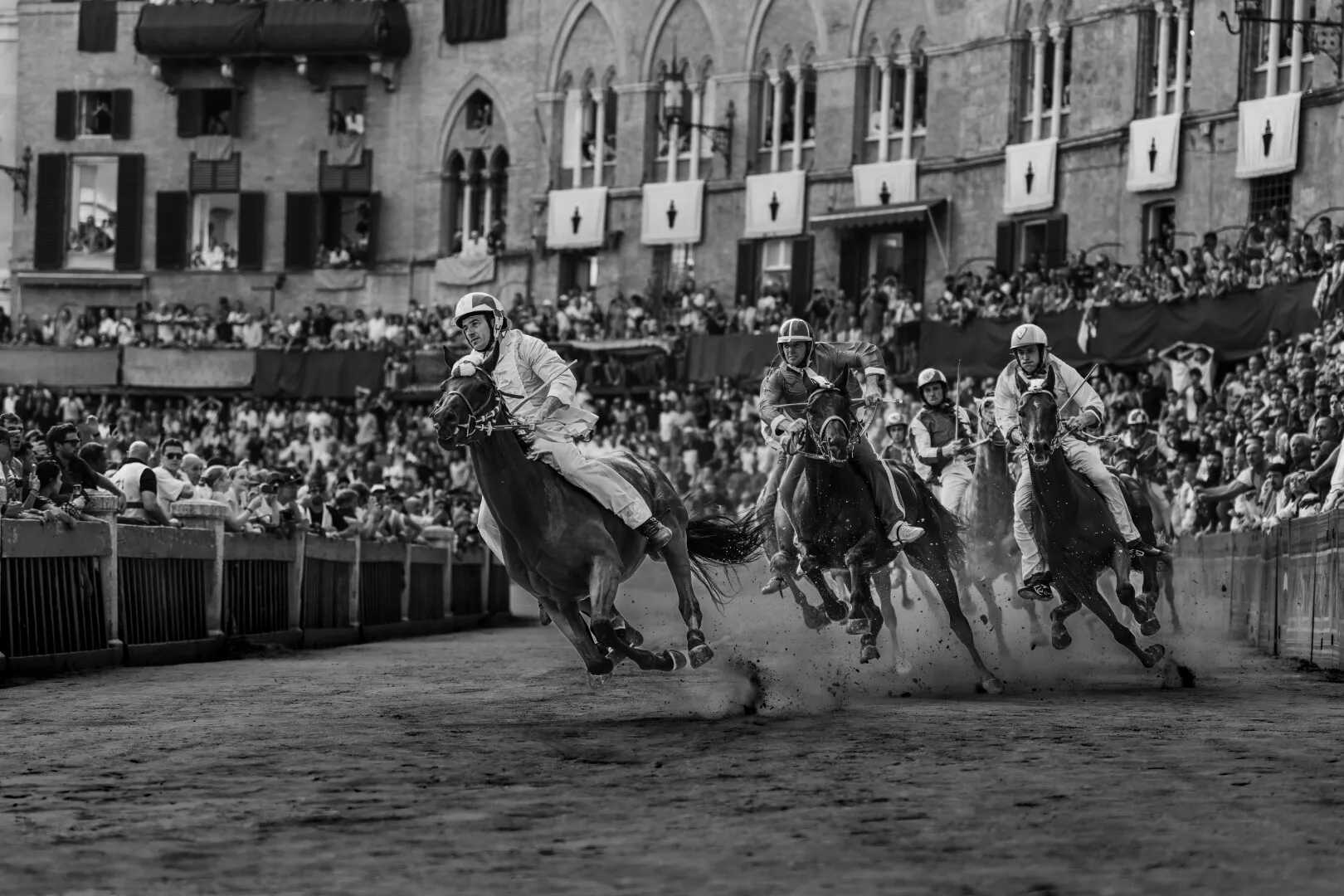 Black and white photo of jockeys riding horses at high speed in a crowded horse race with spectators watching closely.