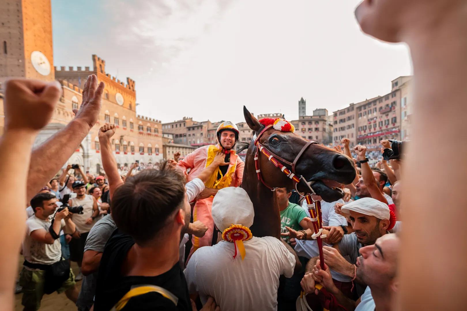 Jockey in pink and yellow gear riding a decorated brown horse, surrounded by cheering crowd in a historic square.