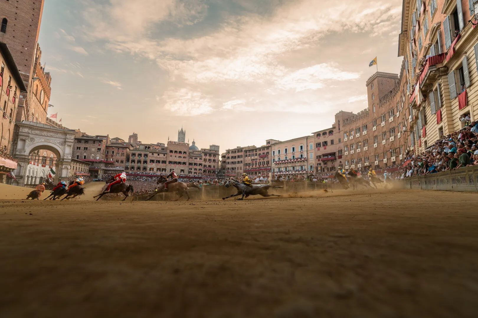 Jockeys riding horses racing around a dirt track in a historic city square with crowd-filled balconies and buildings.