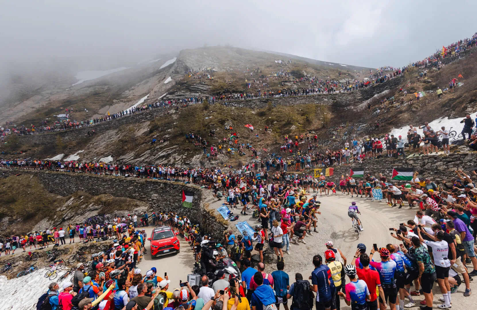 Cyclist racing uphill on a winding mountain road surrounded by large crowds of spectators on both sides and on the hillside.