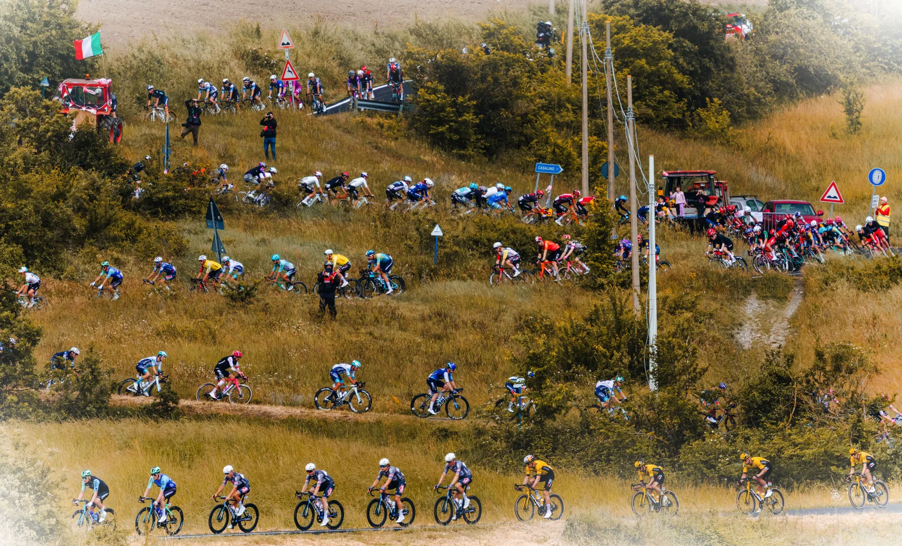 Large group of cyclists riding on a rural dirt road and grassy hillside during a road cycling race, with spectators and vehicles in the background.