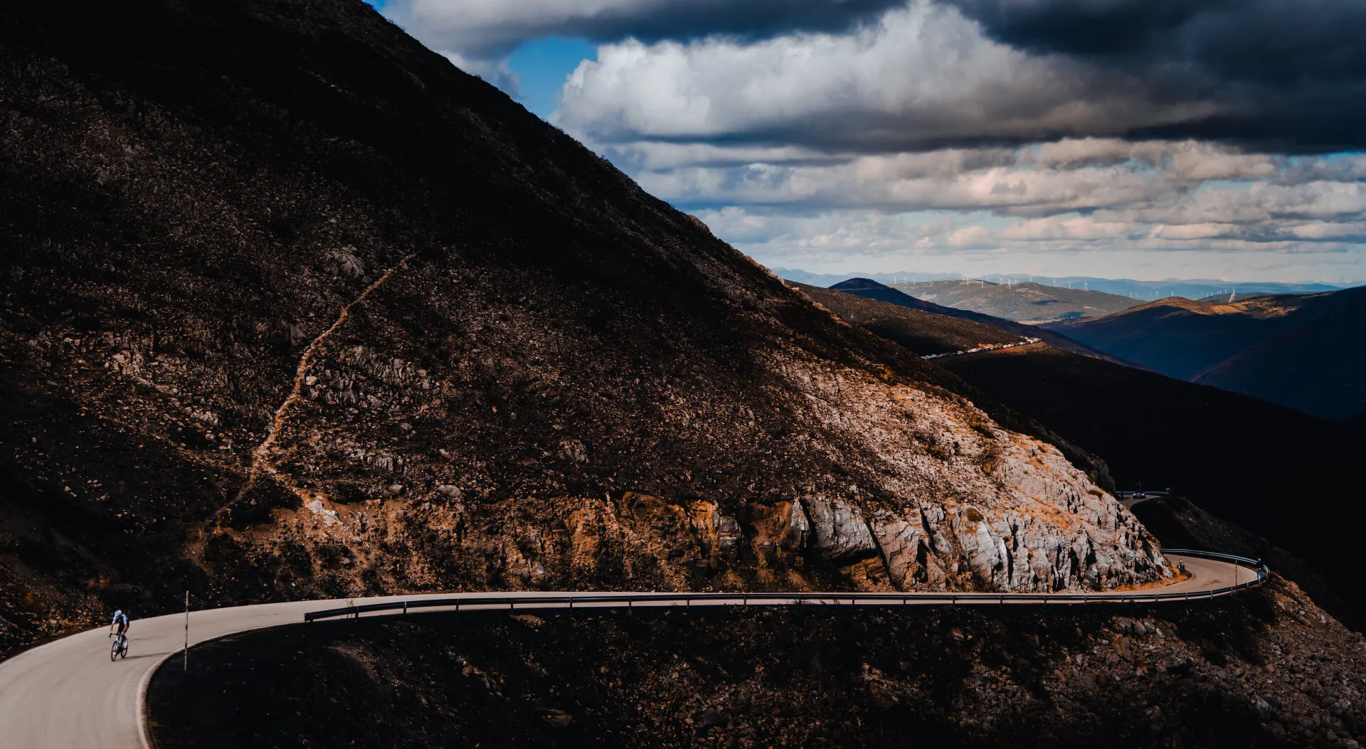 Cyclist riding on a winding mountain road with rocky terrain and cloudy sky.