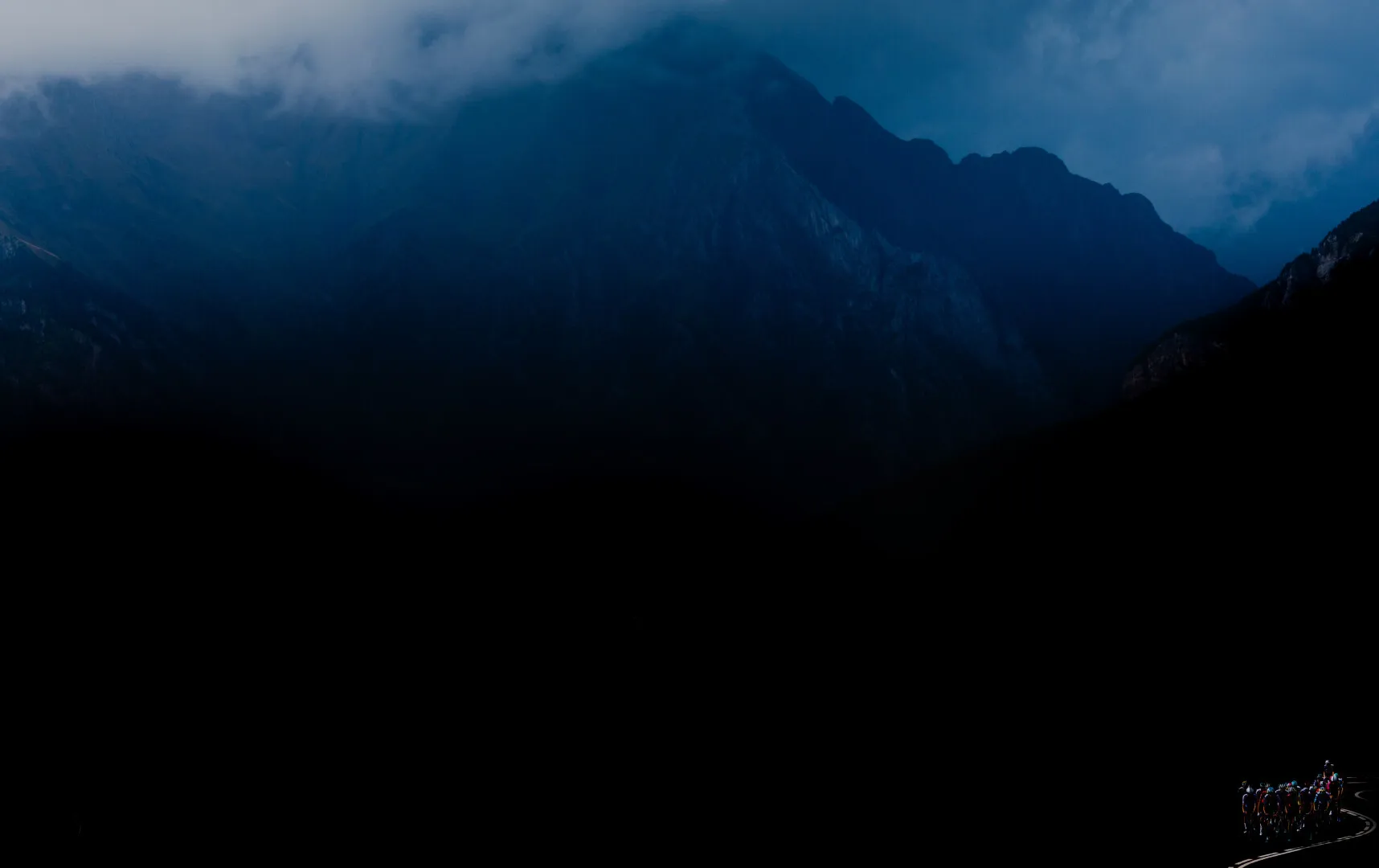 Cyclists riding on a winding road at night with dark mountains and cloudy sky in the background.