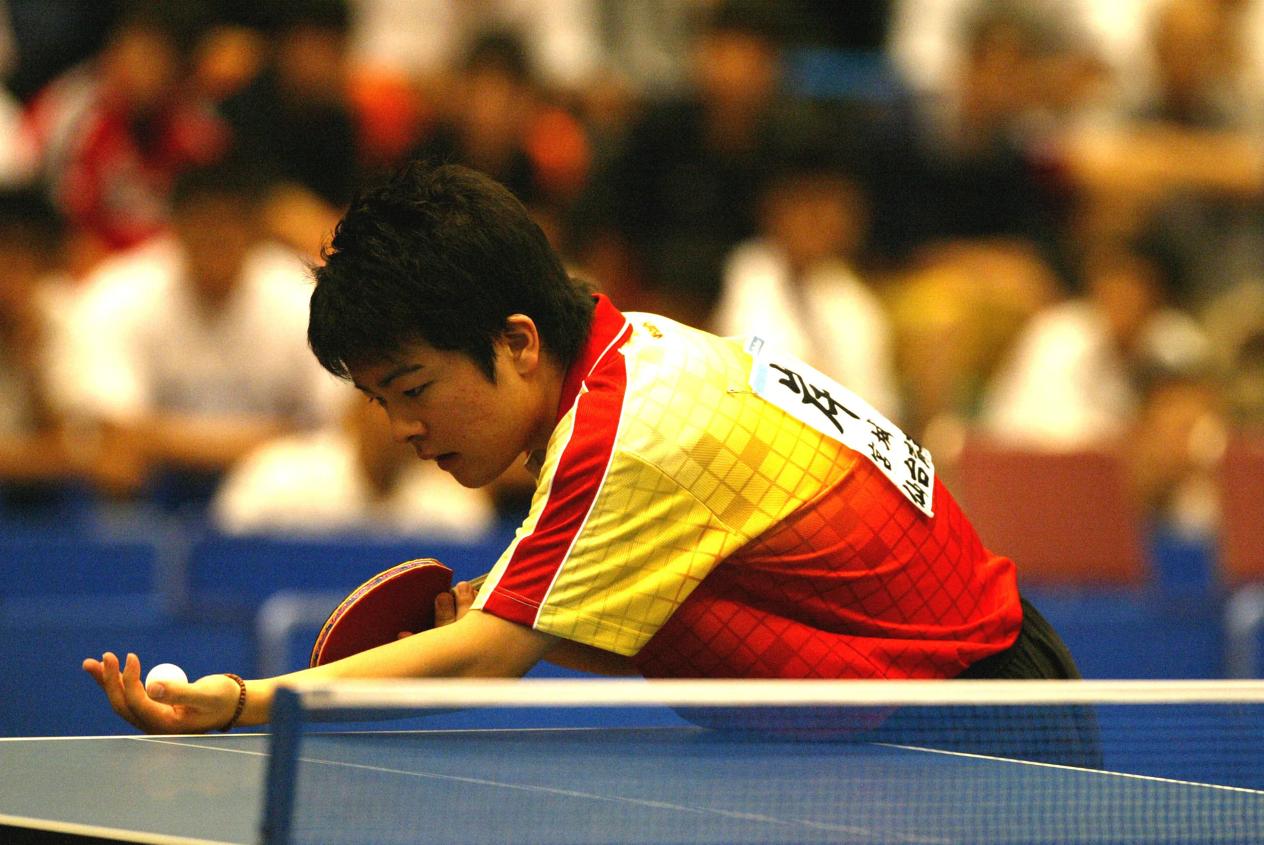 Table tennis player in a red and yellow shirt preparing to serve the ball during a match.