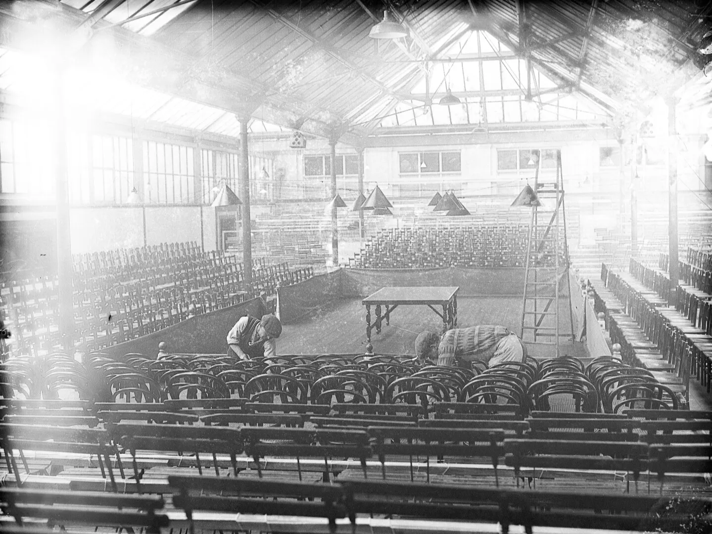 Black and white photo of two boys cleaning chairs in an empty large auditorium with wooden chairs, a table in the center, and a ladder near the stage.