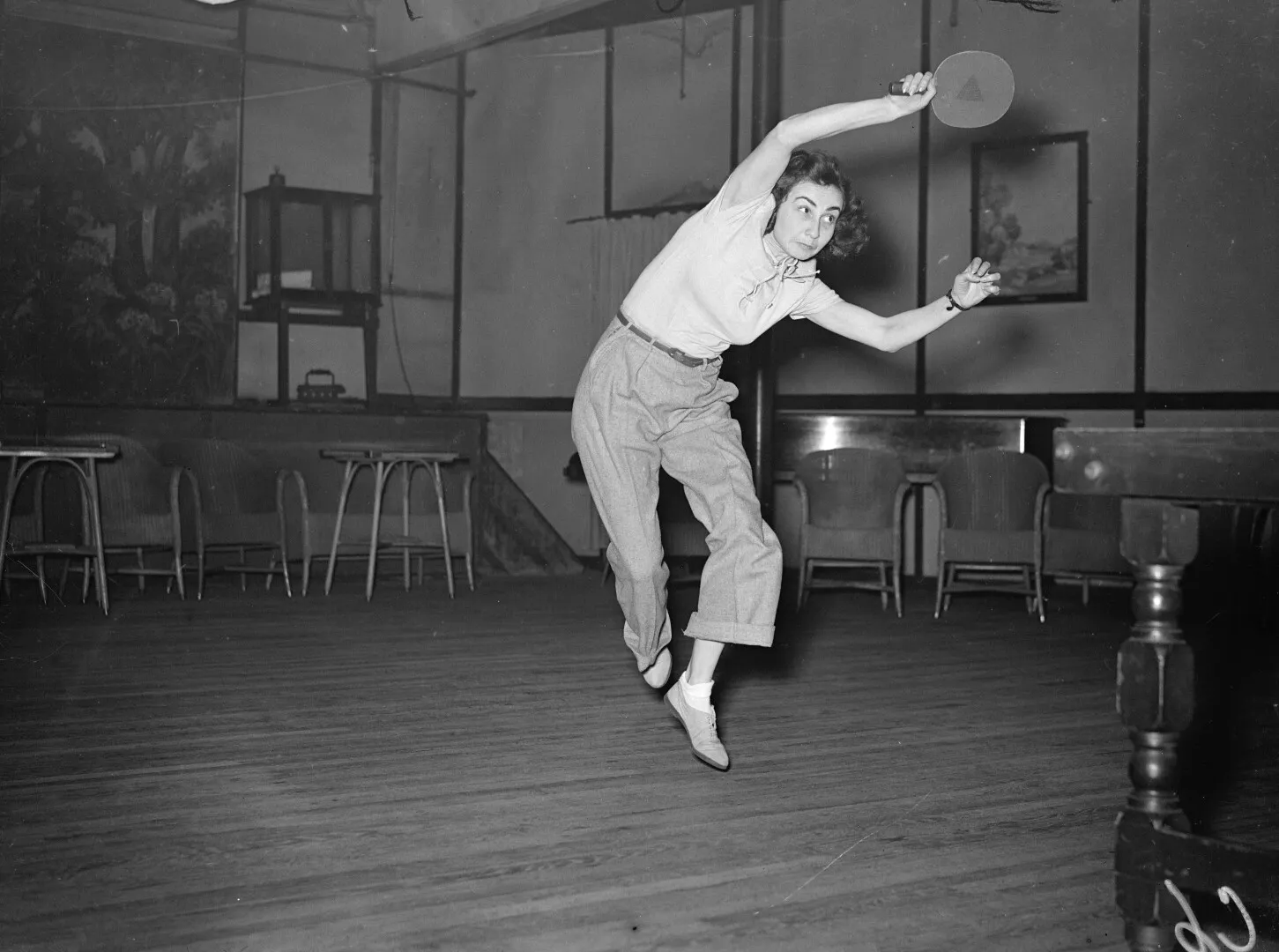 Woman in mid-air playing table tennis indoors, poised to hit the ball with a paddle.