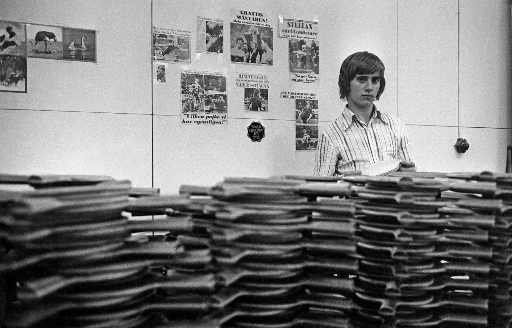 Young man in a striped shirt standing behind several tall stacks of papers with newspaper clippings on the wall behind him.