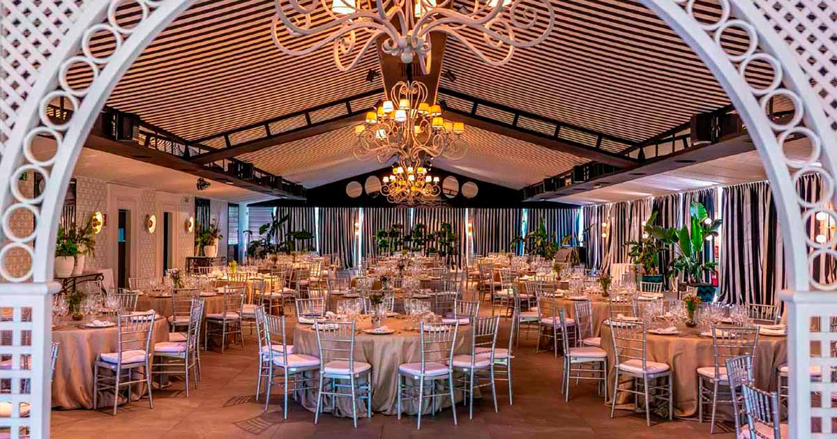 Salón Colonial de Club de Tiro Madrid con mesas redondas vestidas con manteles beige, sillas plateadas y candelabros colgantes decorativos.