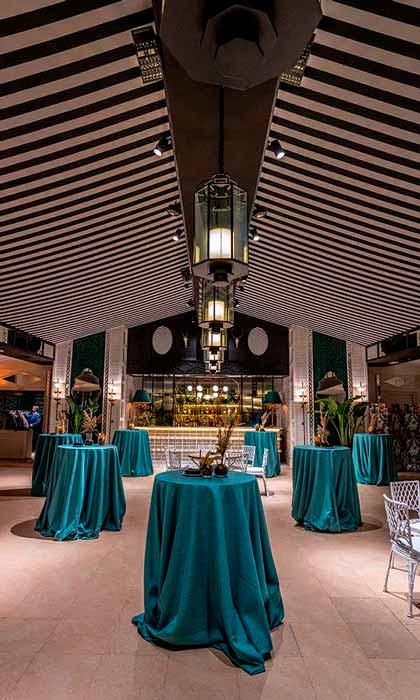 Interior colonial elegante con mesas altas cubiertas de manteles verdes y luminarias colgantes bajo un techo con vigas oscuras.