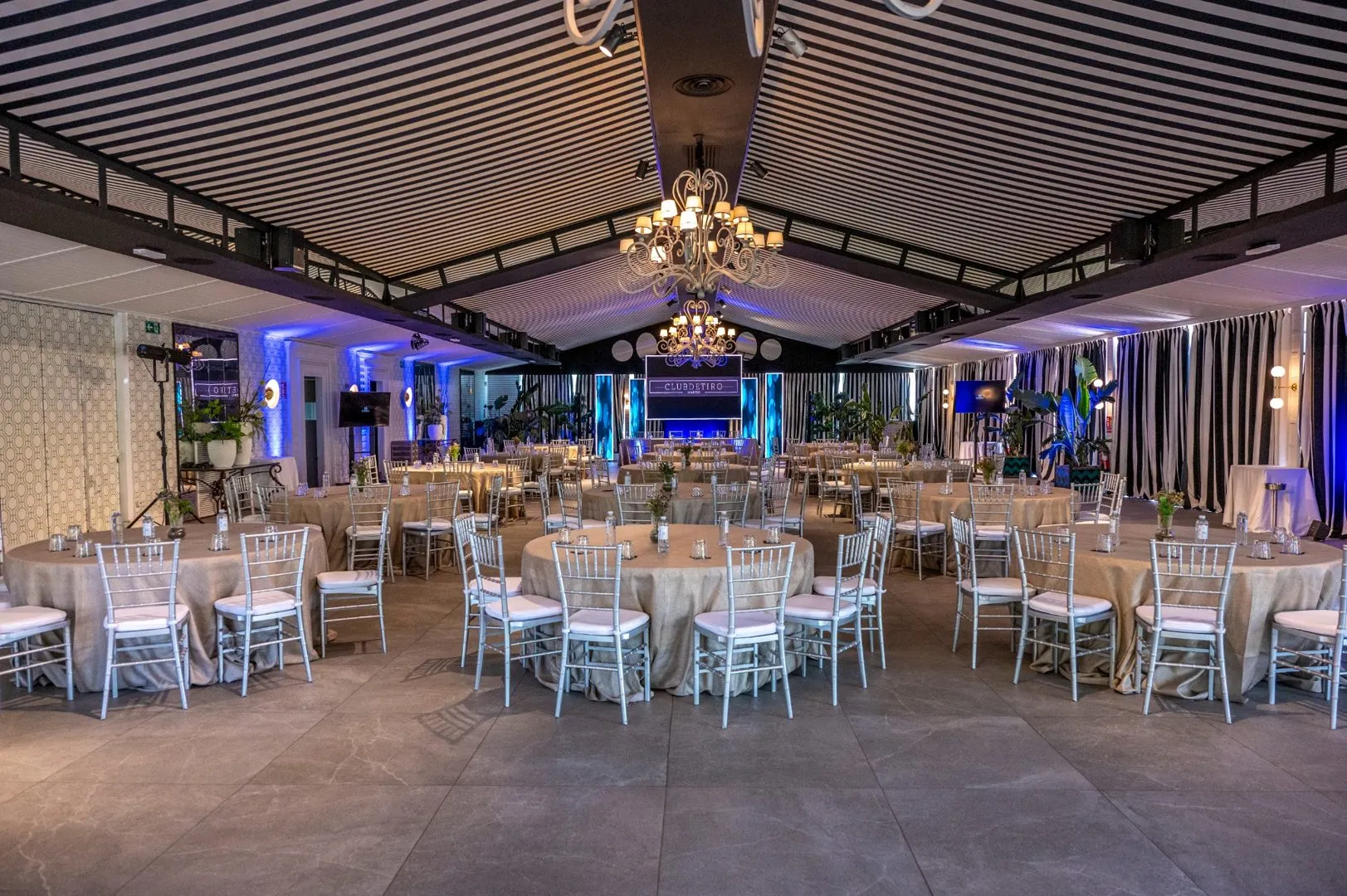 Salón Colonial de Club de Tiro Madrid con mesas redondas vestidas con manteles beige, sillas plateadas y candelabros colgantes decorativos.