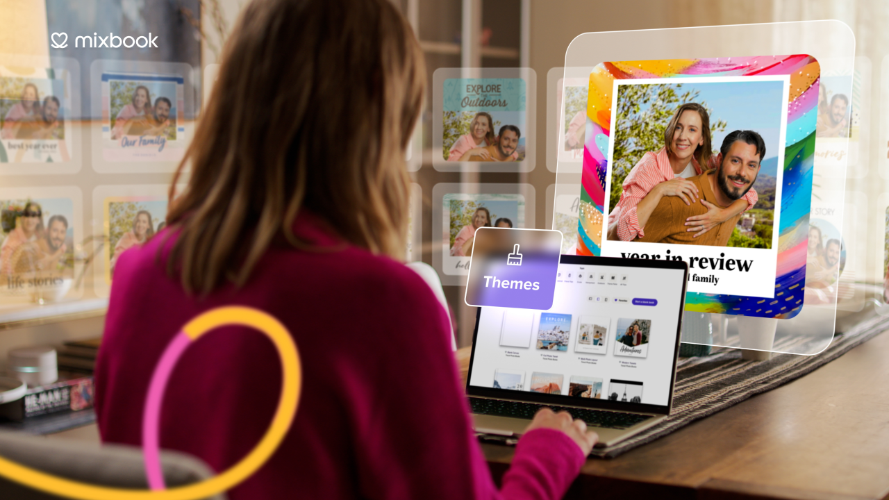 Person in a magenta sweater using a laptop with photo book themes on screen, with floating images and a colorful photo of a couple titled 'year in review' in the background.