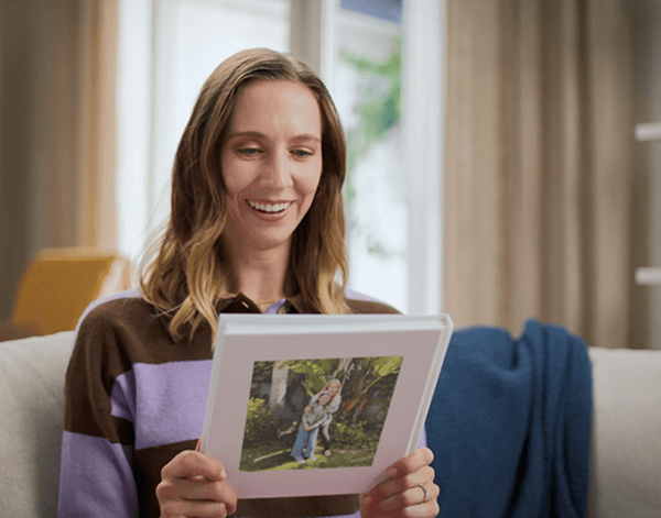 Smiling woman sitting on a couch holding and looking at a framed photo with a picture of children outdoors.