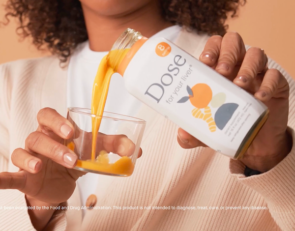 Person with curly hair pouring orange Dose juice into a clear glass cup against a peach background.