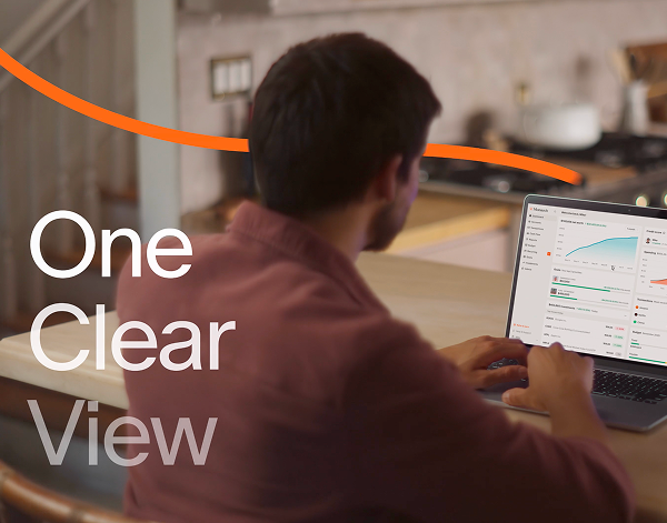 Man seated at a wooden table working on a laptop displaying dashboards with graphs and analytics in a kitchen setting, with overlay text 'One Clear View'.