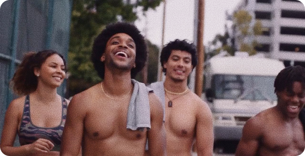 Four young adults smiling and walking outdoors, three men are shirtless, one woman wears a sports bra.