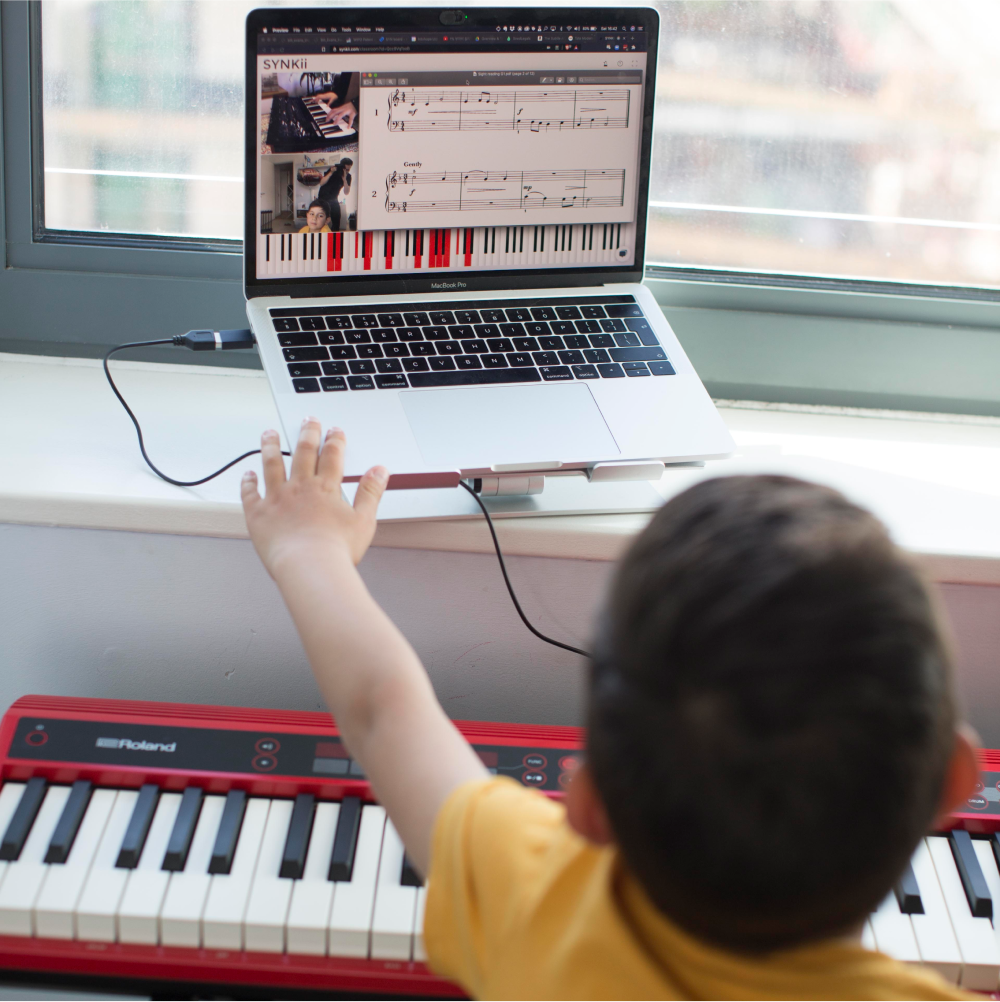 Child reaching toward a laptop displaying SYNKii classroom and keyboard app, with displayed notes played by the teacher