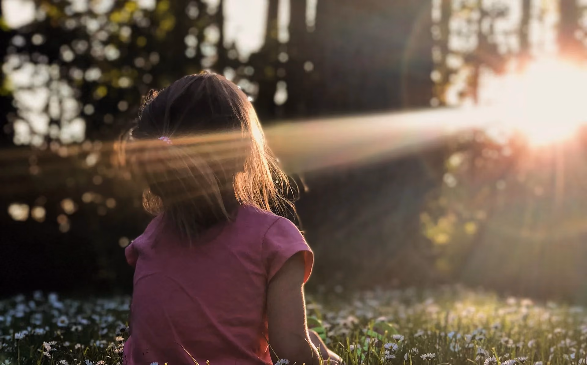 A little girl sitting in the grass looking at the sun.