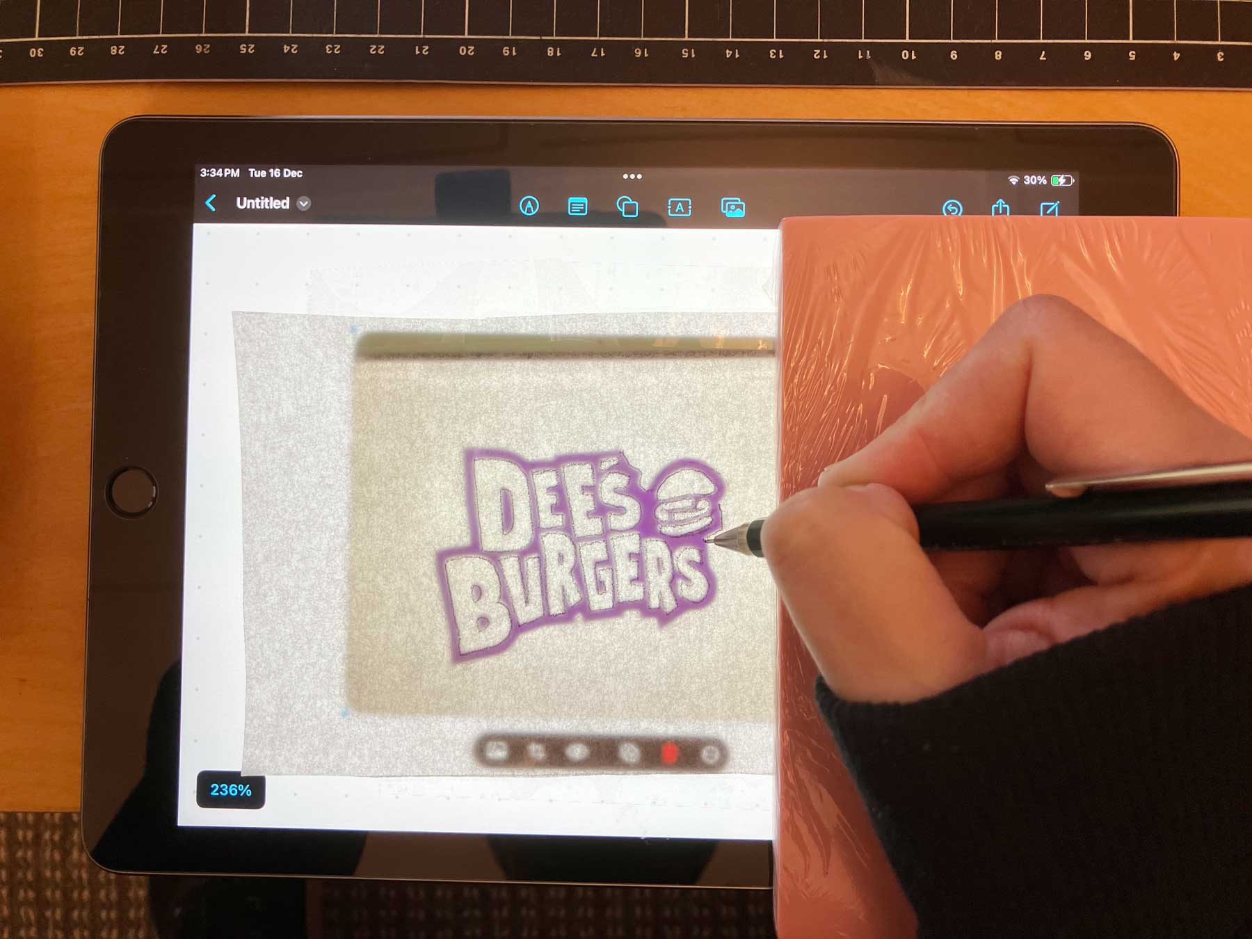 Hand tracing the Dee's Burgers logo on an iPad screen using a pencil and tracing paper