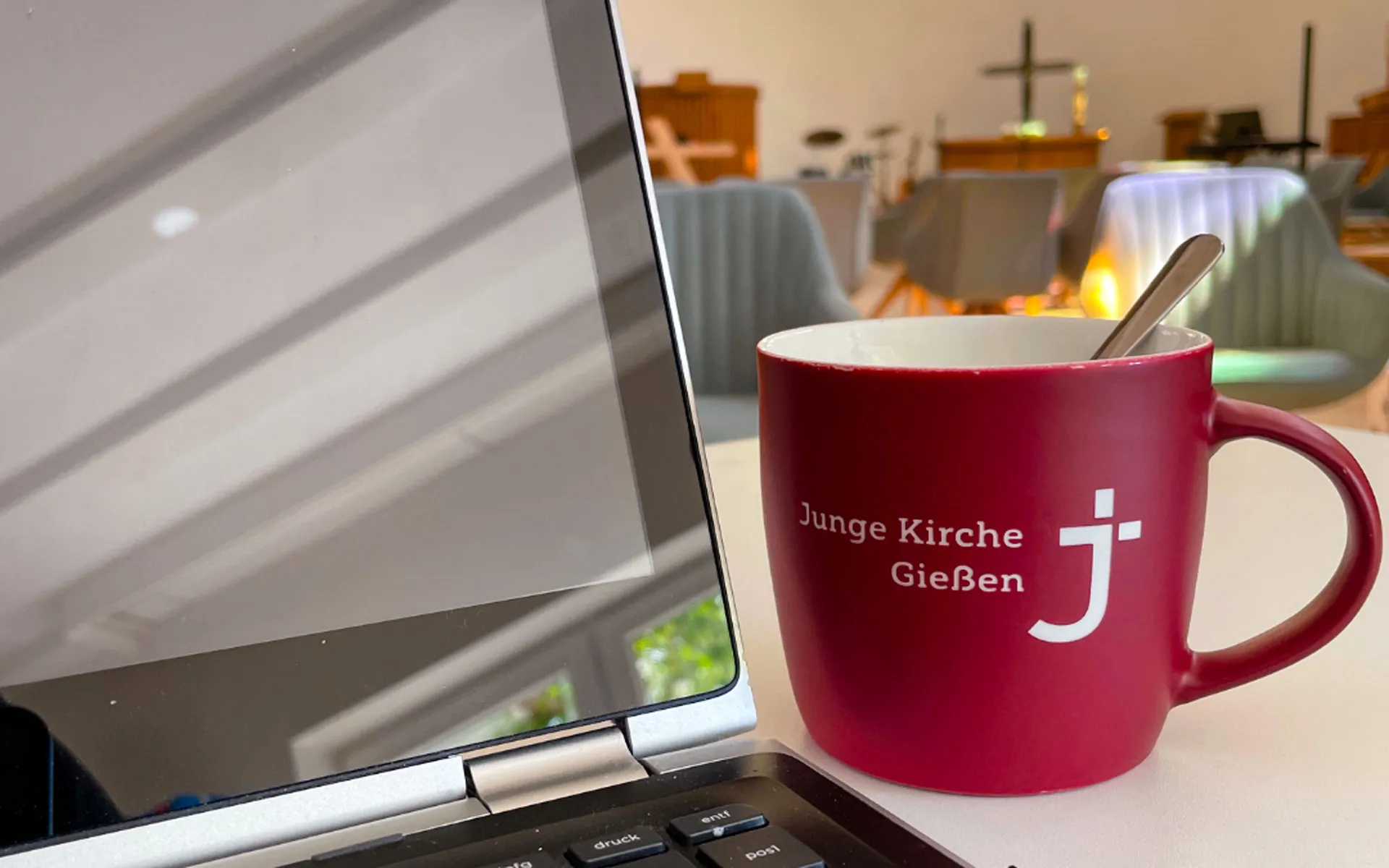 JuKi-Tasse neben Laptop; im Hintergrund Blick auf Altar.