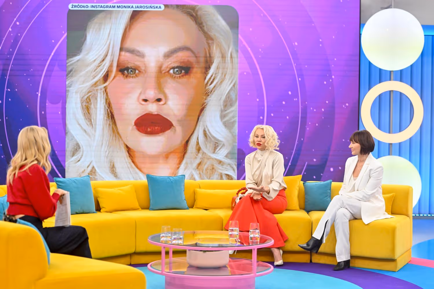 Ewa Abramowicz siedzi na żółtej kanapie w telewizyjnym studiu, obok dwóch kobiet, z dużym ekranem w tle pokazującym portret kobiety z blond włosami i czerwonymi ustami.