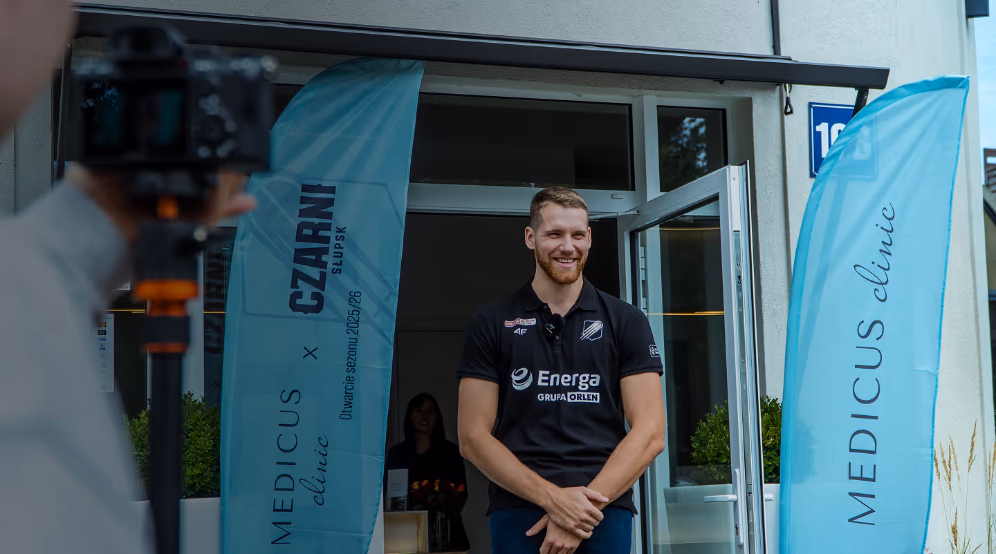 Mężczyzna w czarnej koszulce stoi uśmiechnięty przy otwartych drzwiach kliniki Medicus obok niebieskich flag z napisami.