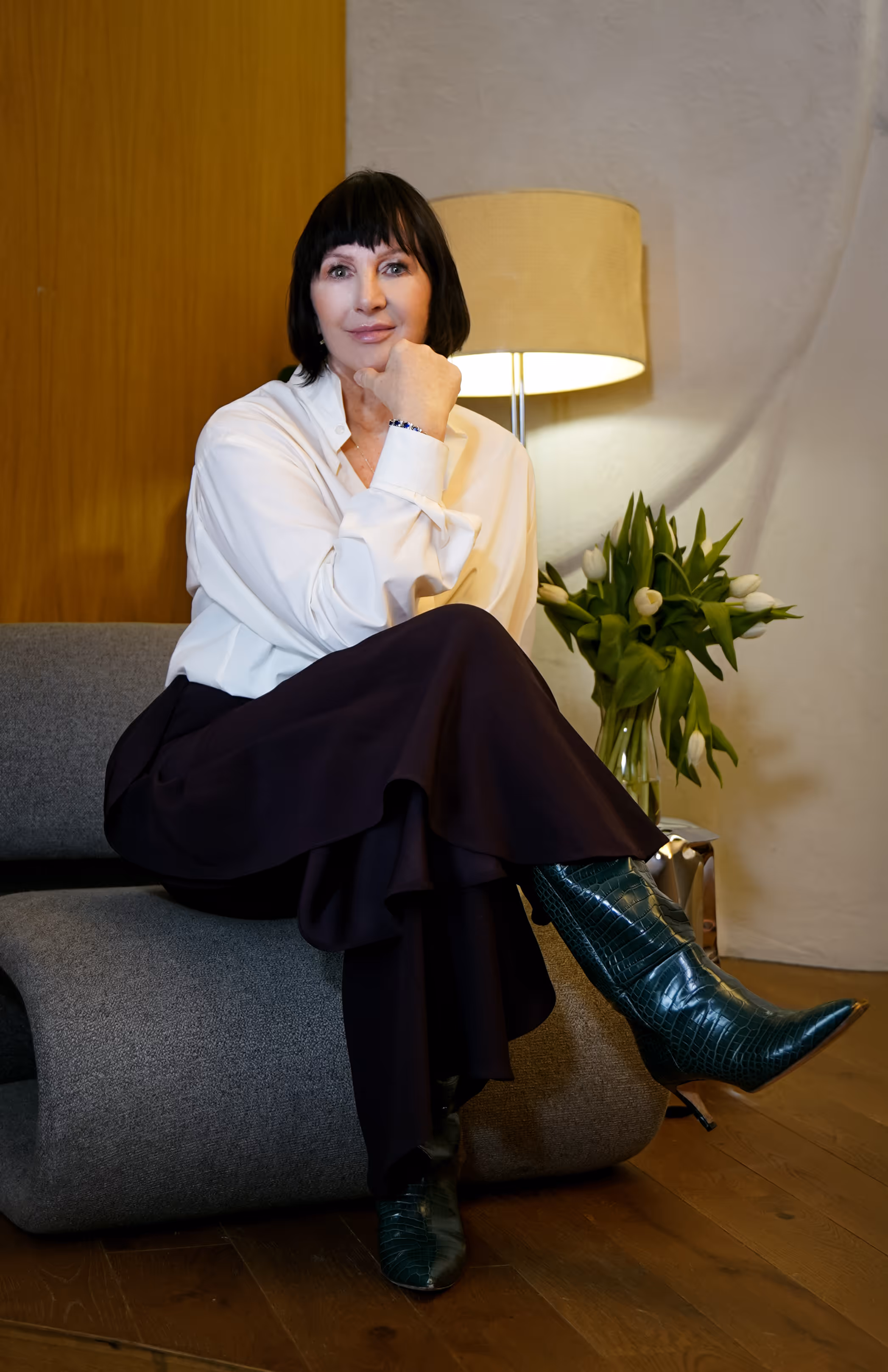Ewa Abramowicz w białej koszuli, czarnej spódnicy i zielonych skórzanych botkach siedzi na szarej sofie z ręką pod brodą, w tle lampa i wazon z białymi tulipanami.