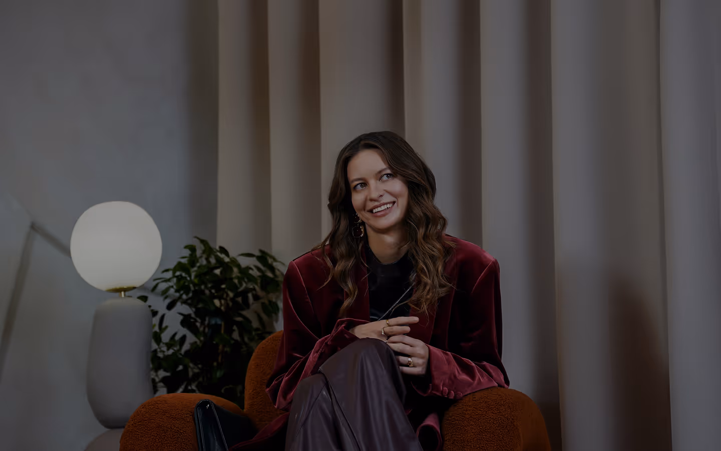 Uśmiechnięta kobieta z długimi falującymi włosami w bordowej marynarce siedzi na brązowym fotelu w nowoczesnym wnętrzu.