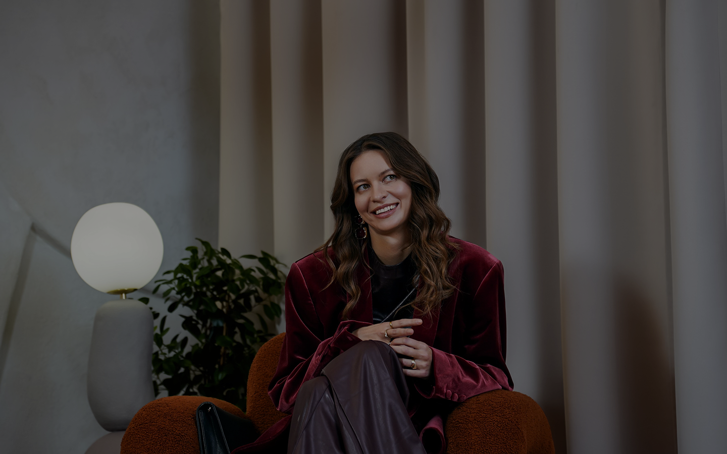 Uśmiechnięta kobieta z długimi falującymi włosami w bordowej marynarce siedzi na brązowym fotelu w nowoczesnym wnętrzu.