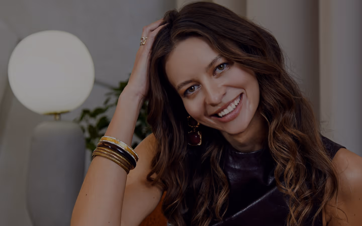 Uśmiechnięta młoda kobieta z długimi falującymi włosami w bordowej marynarce siedzi na pomarańczowym fotelu przy lampie i zielonej roślinie.