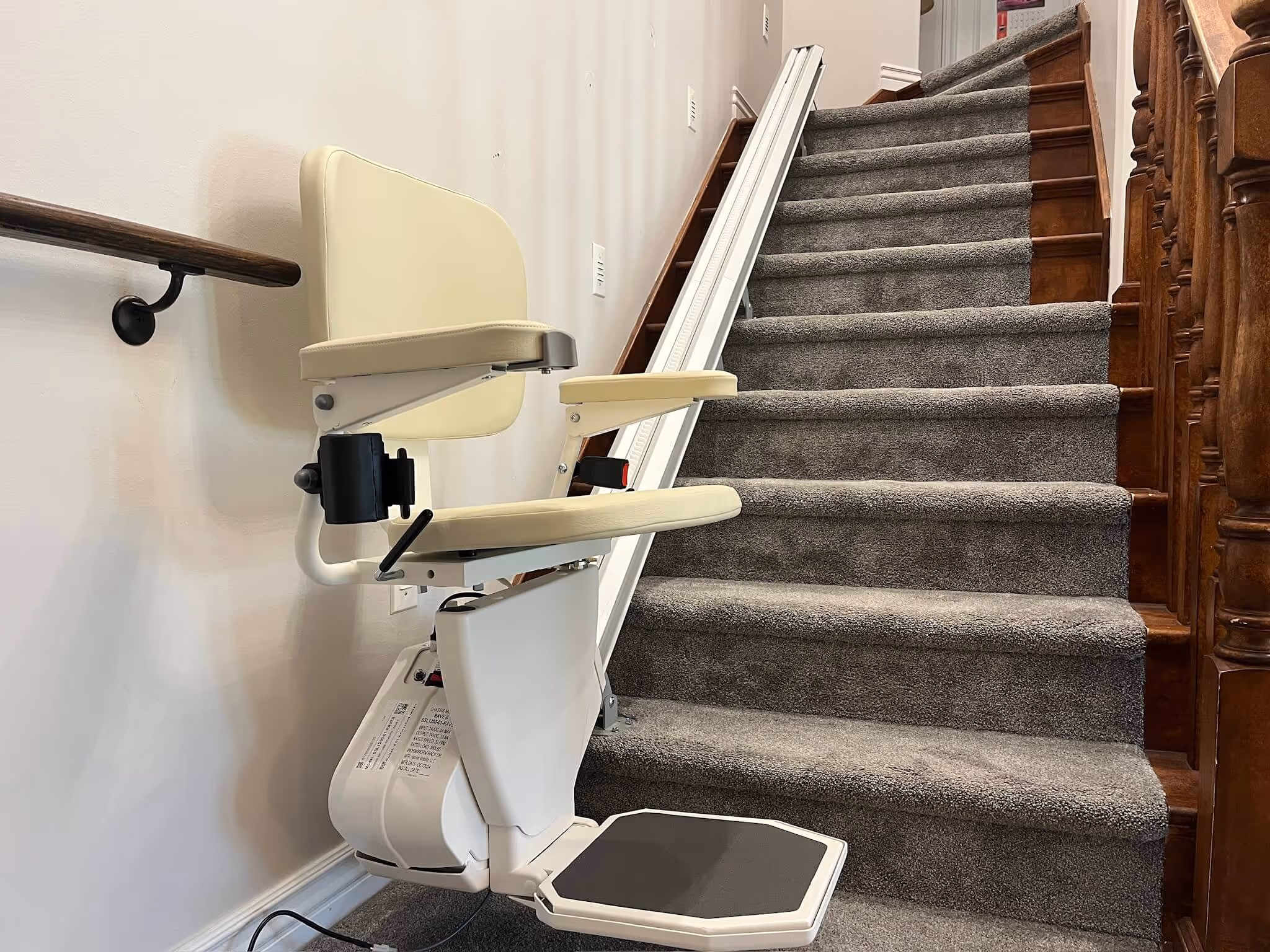 A white and tan stair lift chair parked at the bottom of a carpeted wooden staircase, with its rail running alongside the steps.