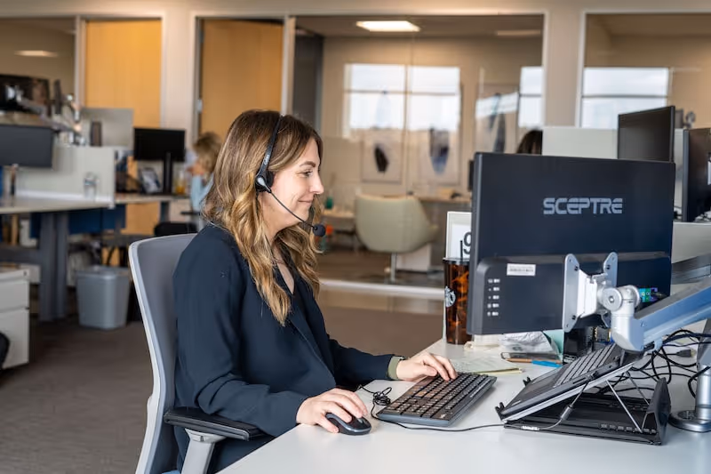 A woman sitting at a desk in an office with a headset, working at a desktop computer
