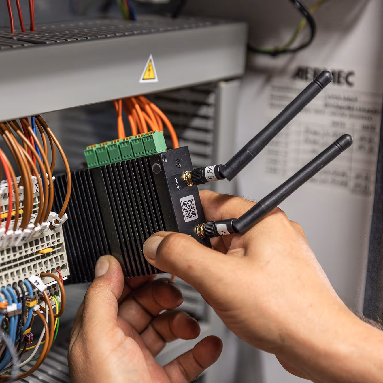 Person holding black wireless module with two antennas and connecting to industrial equipment with wires.