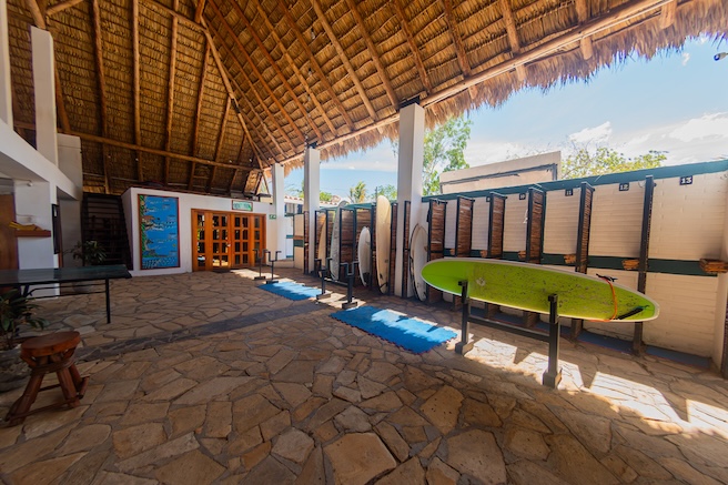 Surfboard storage area at Miramar SurfCamp in Nicaragua, featuring boards on racks under a large thatched roof with stone flooring.