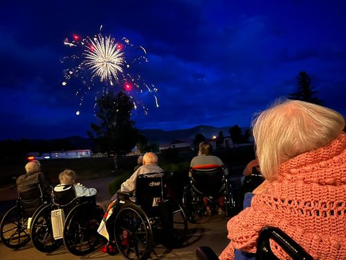 Residents at Pine Ridge Rehabilitation and Healthcare Center gather outdoors, smiling and watching fireworks light up the night sky on July 4th, expressing joy and camaraderie.