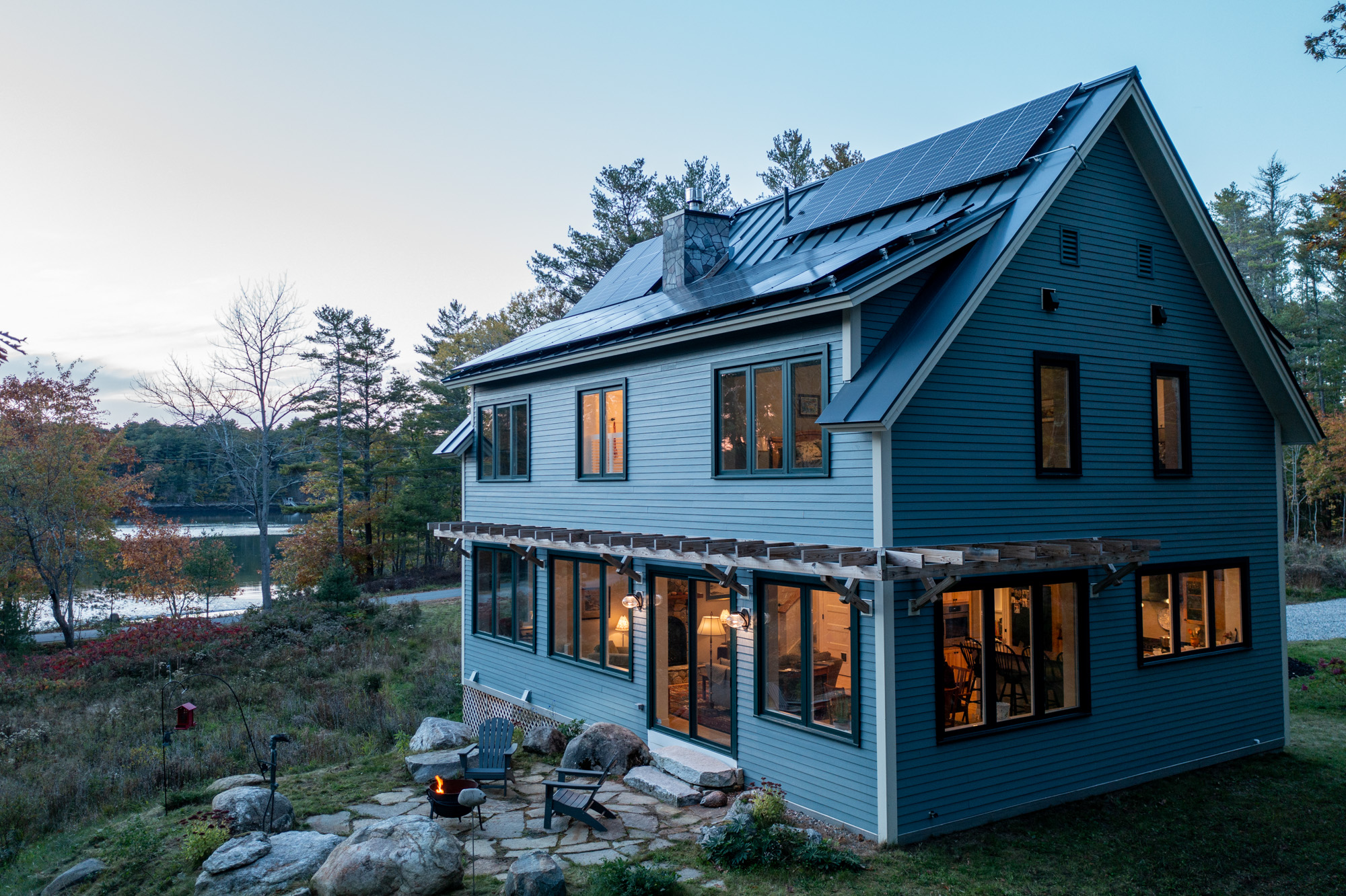 Blue solar-powered house with large windows near lake and forest at dusk