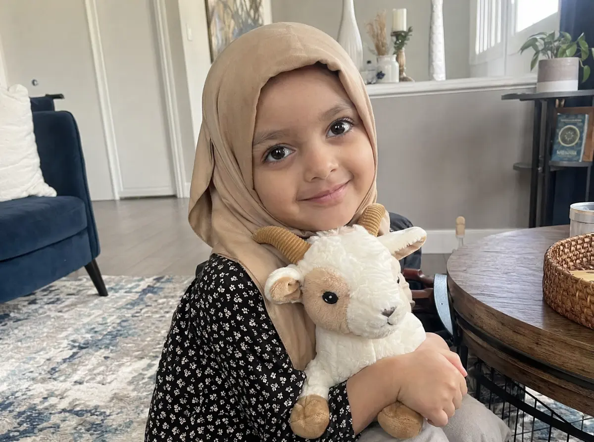 A young girl wearing a beige headscarf and black floral dress smiles warmly, hugging a plush goat toy, sitting in a cozy, well-lit living room.