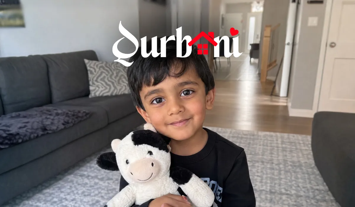 Young child smiling warmly while holding a plush cow toy, sitting in a cozy living room. The mood is cheerful and welcoming. The word “Qurbani” is stylized at the top.