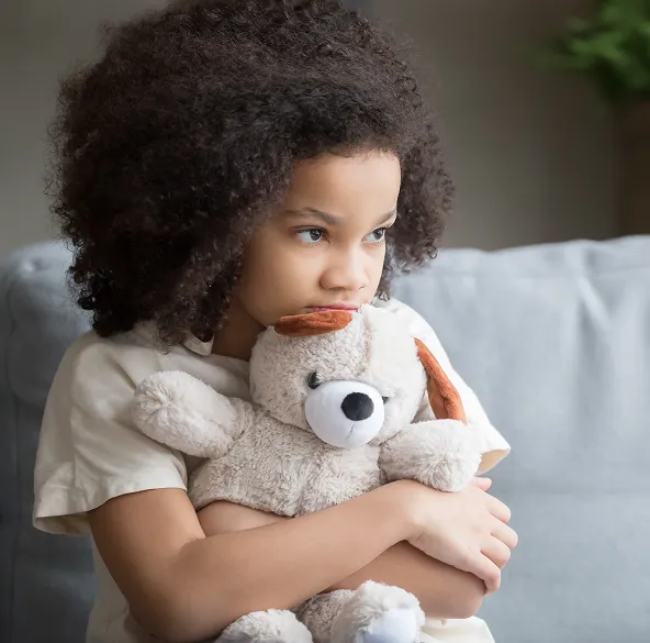 A young child with curly hair sitting on a couch, holding a stuffed animal and looking thoughtful or sad.