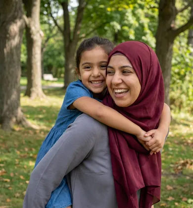 A woman wearing a maroon hijab gives a piggyback ride to a young girl in a blue dress, both smiling joyfully while outdoors in a wooded park.