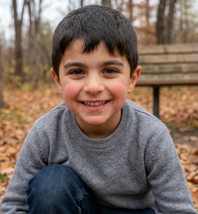 A young boy with dark hair and rosy cheeks smiling at the camera, wearing a gray sweater, crouched in a park with fallen autumn leaves and a wooden bench in the background.