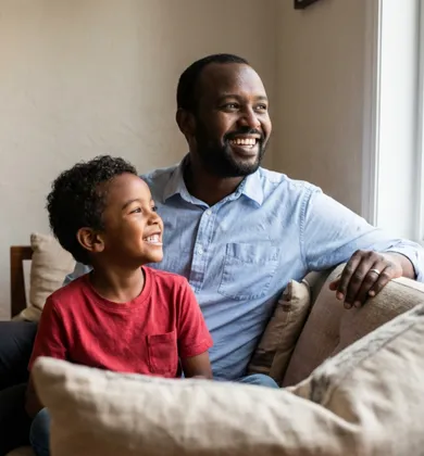 A Black father and his young son sitting on a couch together, both smiling and looking out a window.
