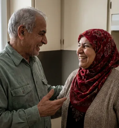 An elderly man with grey hair wearing a green shirt laughs while talking to a smiling elderly woman wearing a patterned red hijab and a beige cardigan in a home kitchen.