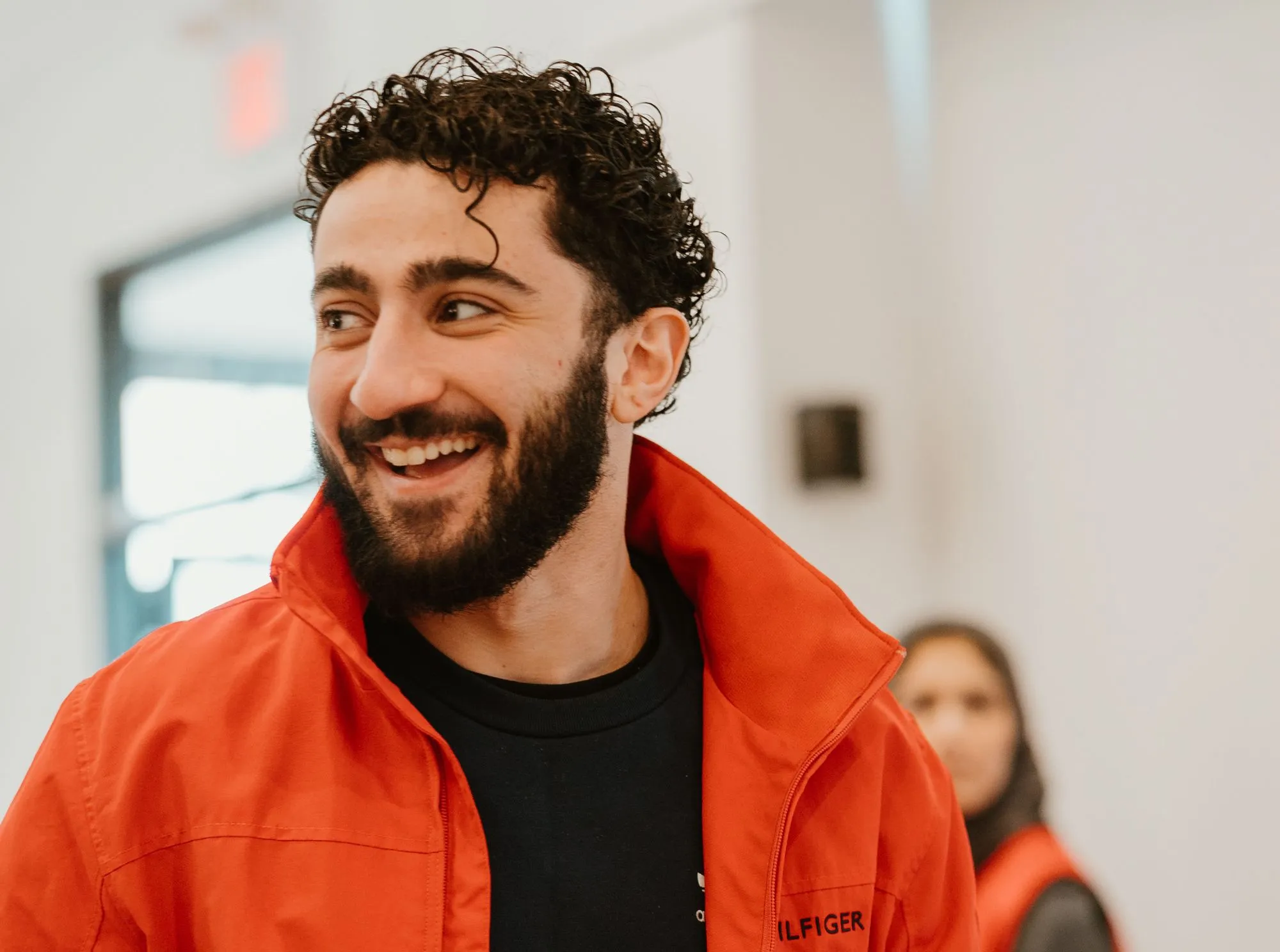A smiling man with curly dark hair and a beard, wearing an unzipped red Tommy Hilfiger jacket over a black shirt, looking toward the left.