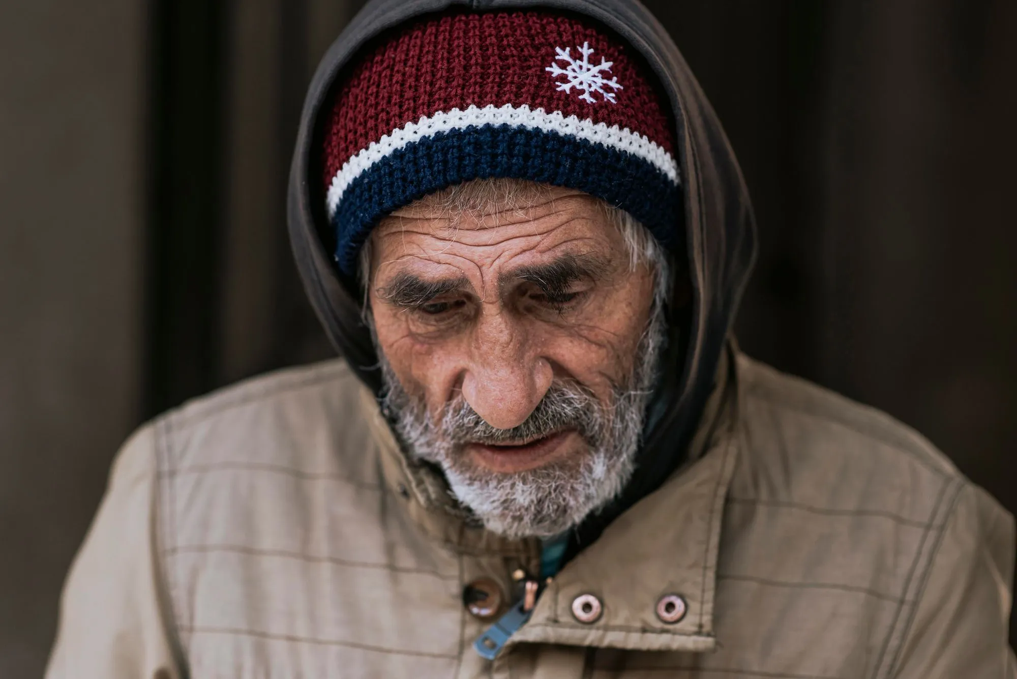 Close-up portrait of an elderly man with deep facial wrinkles and a grey beard, wearing a brown jacket with the hood up over a maroon, white, and navy blue knit beanie featuring a white snowflake design.