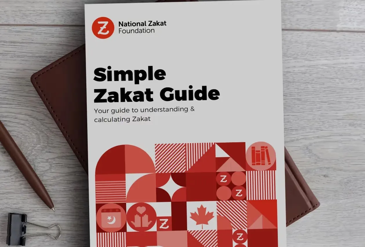 The cover of a Simple Zakat Guide booklet by the National Zakāt Foundation, featuring geometric red and white patterns, a pen, and a binder clip on a wooden surface.