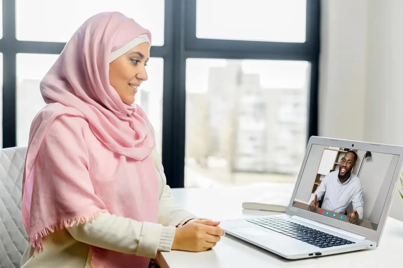 A woman wearing a pink hijab sits at a desk, smiling while looking at her laptop screen during a video call with a smiling Black man in a white shirt.